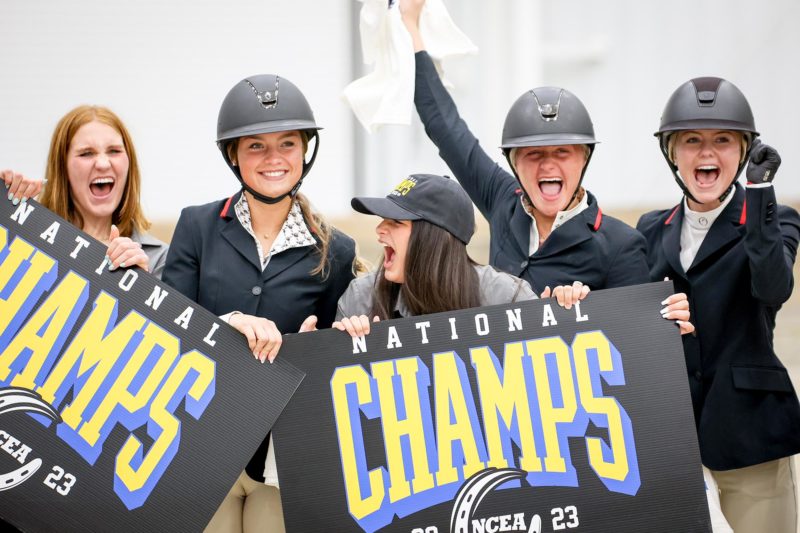 Five girls in equestrian outfits cheer and hold up signs reading "National Champs"