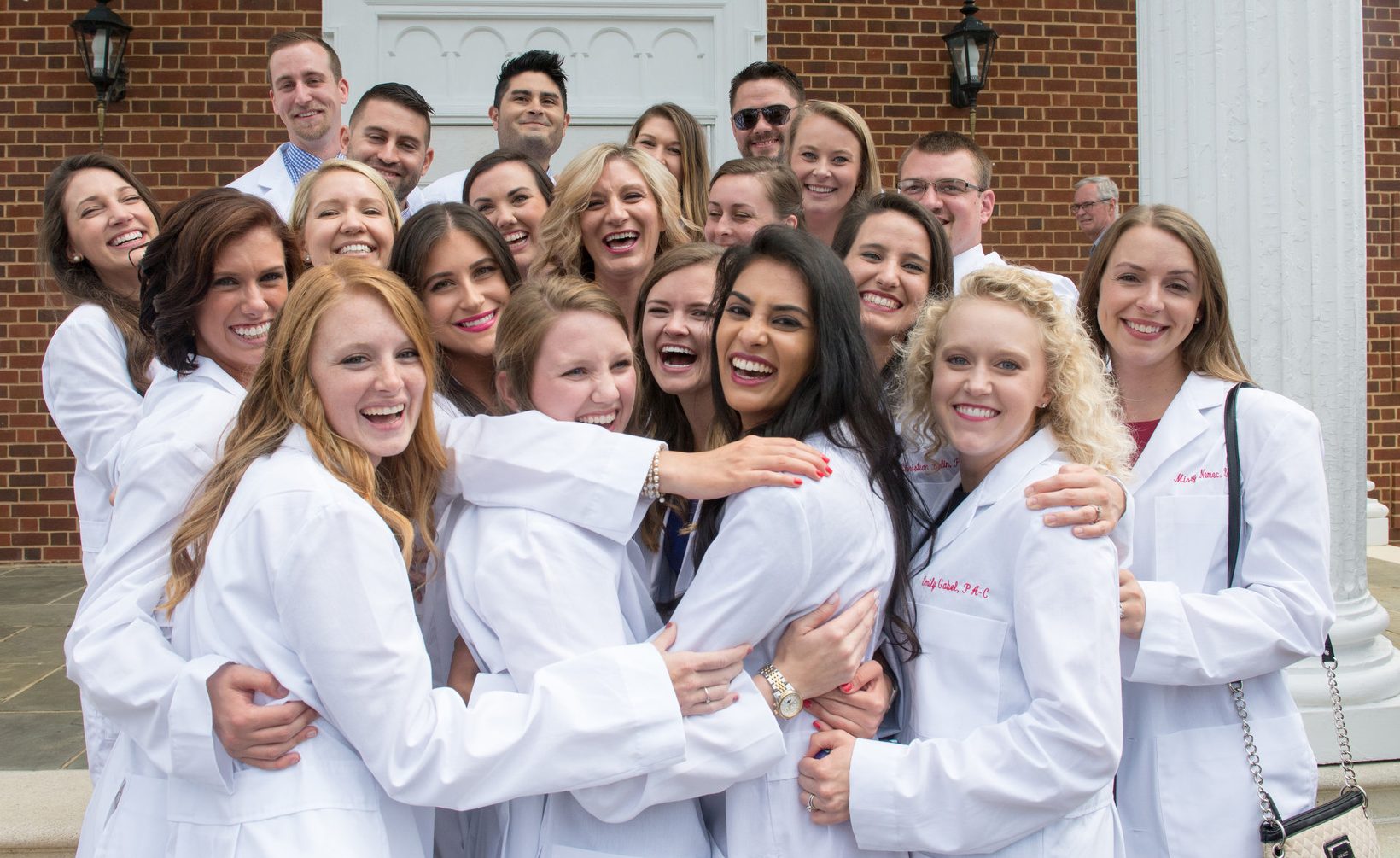 A group of more than 20 students wearing white coats are gathered on the front porch of Snidow Chapel.