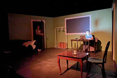 A stage is shown set up as a room; a man sits at a desk while a woman enters through a door.