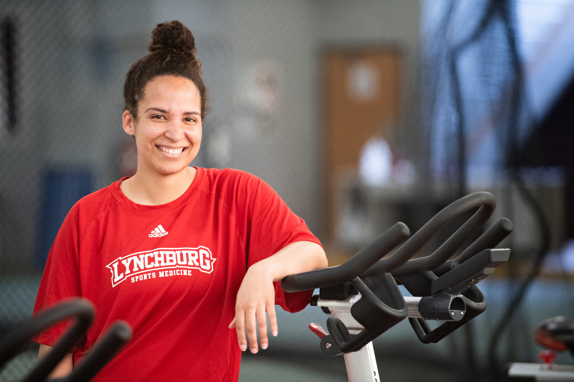 A biracial female student in a red Lynchburg T-shirt, smiling, leaning against an exercise machine
