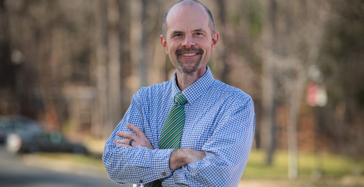 Portrait of a middle-aged white man in a blue plaid button-up and green tie with beard smiling and crossing his arms