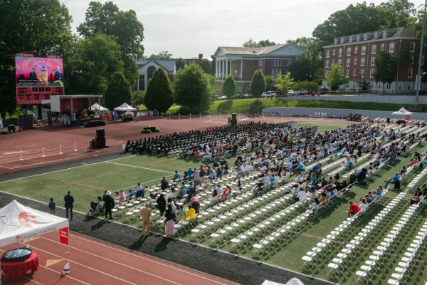 Ceremony on athletic field with white chairs and buildings in the background, blue sky