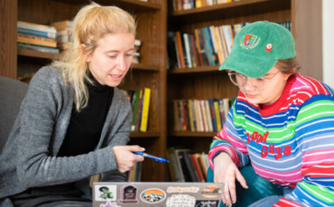 A white woman with long blonde hair dressed in gray and black sits with her laptop covered in stickers and a white female student in a rainbow sweater and baseball hat