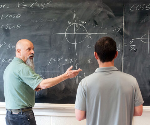 A professor explaining a mathematics problem to a student while standing in front of a blackboard