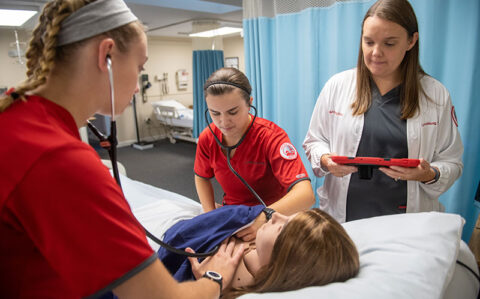 Two nursing students practice on a medical dummy while an instructor looks on.