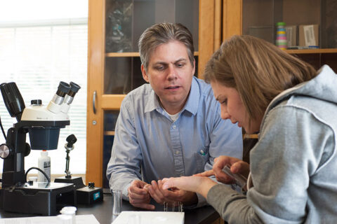 An instructor sits with a student near a microscope.