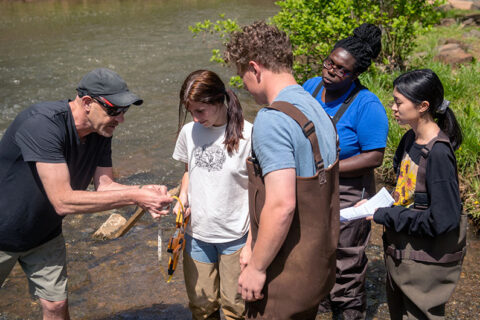A professor is showing four of his students how to use a measuring device. They are standing at the edge of a shallow river.