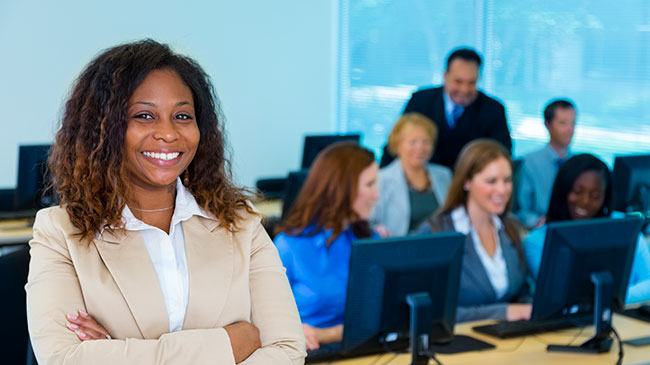A woman stands with her arms crossed with a classroom behind her.