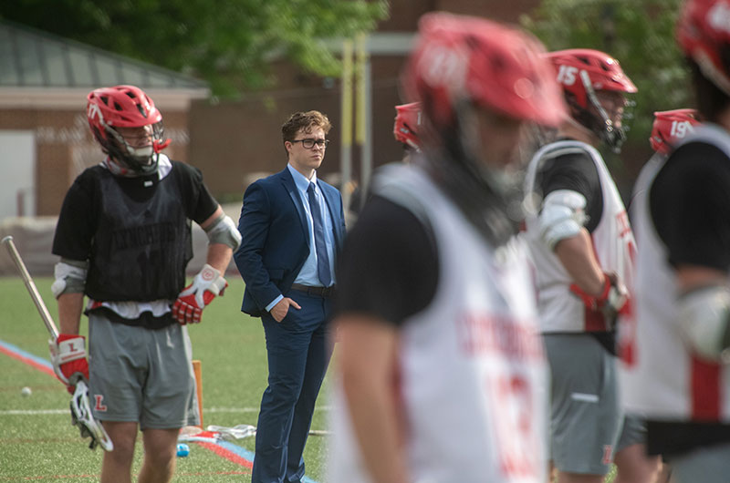 A man in a suit stands on the sidelines of a lacrosse game.