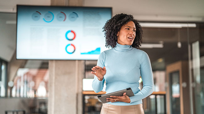 A woman stands at the front of a room in front of a large computer screen