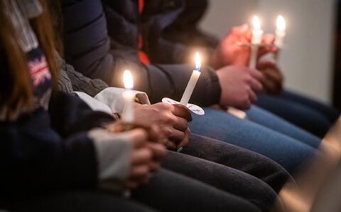 Several people in black and blue jeans and dark coats sit in a pew holding candles