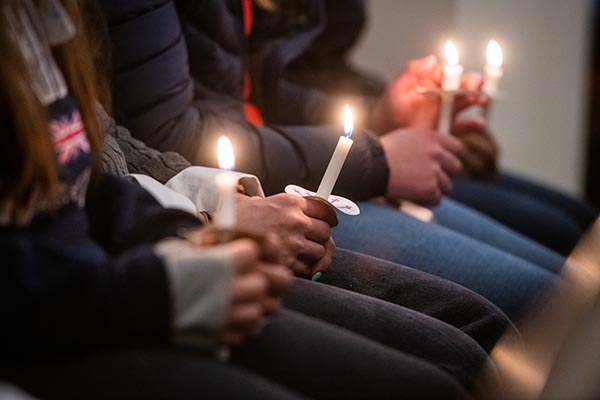 Several people in black and blue jeans and dark coats sit in a pew holding candles