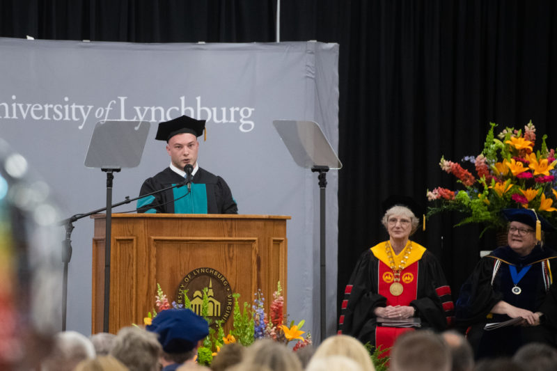 A young white man in academic regalia speaking at a podium with the crowd in the foreground and two middle-aged white women in academic regalia sitting on stage and watching him