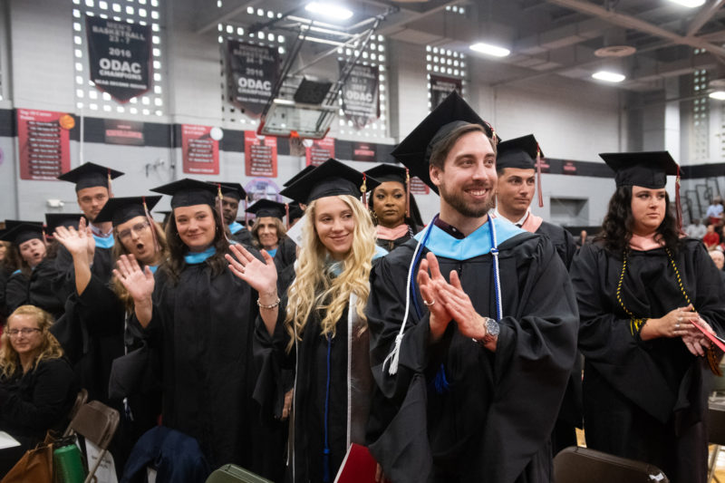 A group of graduate students in academic regalia smiling and applauding