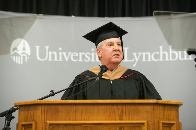 A white man with gray hair in academic regalia speaking at a podium