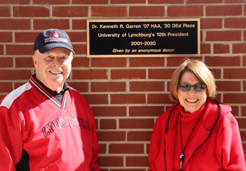 An elderly man with a baseball hat and a red shirt and a middle-aged woman in red, both smiling, in front of a brick wall and plaque
