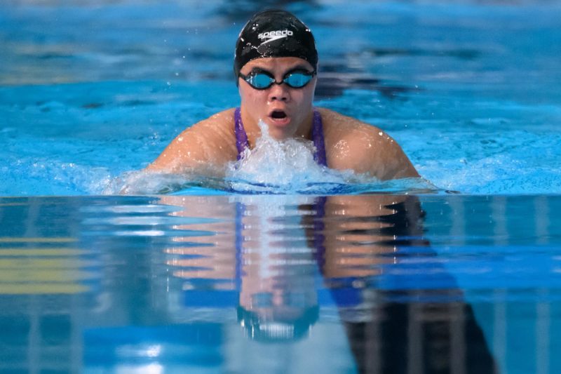 A female swimmer with a black cap and goggles wearing a purple bathing suit breathing as she comes out of the water, her reflection below her