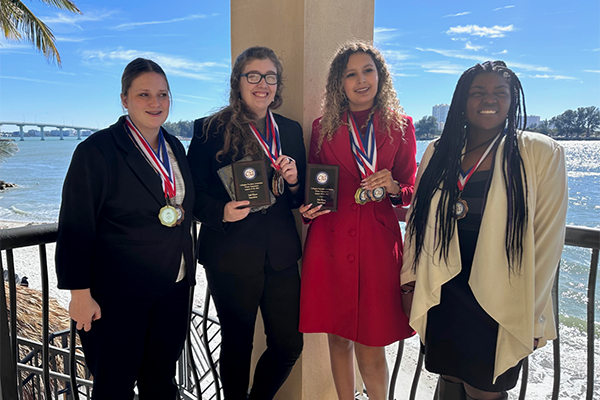 Four young women in black suits and dresses stand outside with a blue sky and palm trees in the background.