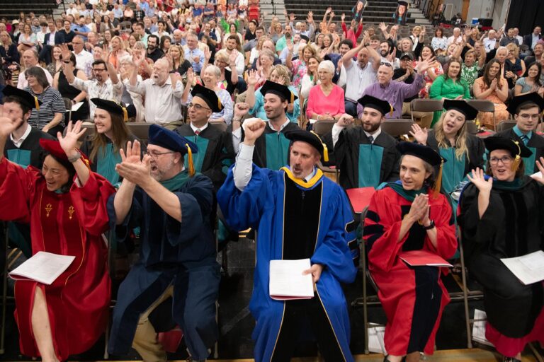 A seated crowd of family, faculty, and graduating students who are cheering and clapping.