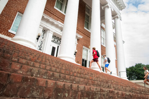 Students walking up the brick steps of a historic building with large white columns, carrying backpacks, on a sunny day with a partially cloudy sky.