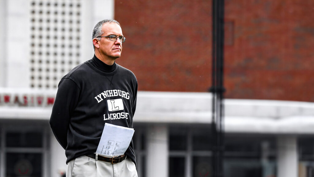 A Lynchburg men's lacrosse coach in a black "Lynchburg Lacrosse" sweatshirt observes the field intently, holding papers, with campus buildings in the background.