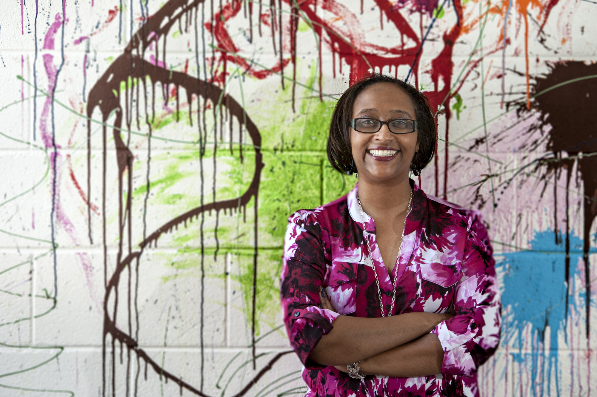 A smiling black woman with dark brown chin length hair stands with her arms crossed. She stands in front of an brightly colored and abstractly painted cinder block wall.