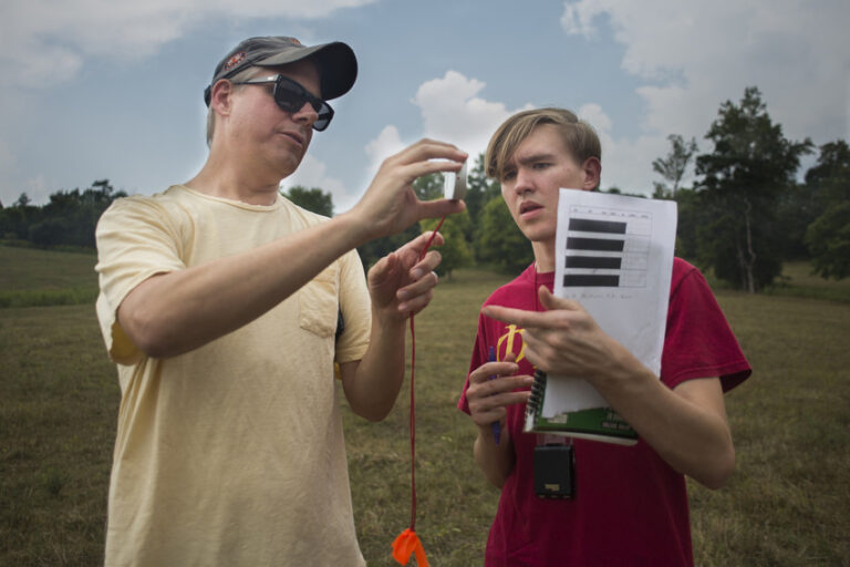 An instructor wearing a cap and sunglasses demonstrates the use of a tool to a student holding a notebook, both standing in a grassy field.