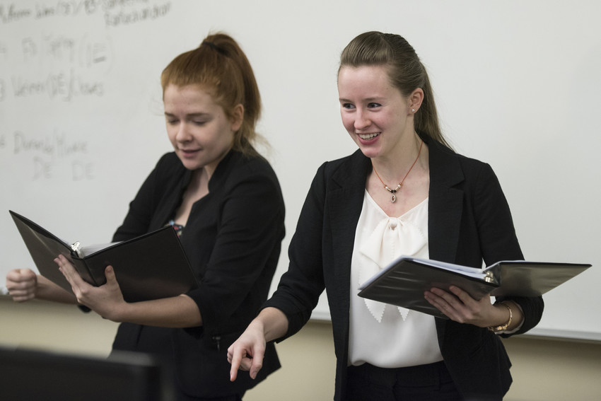 Two students presenting with binders in hand, smiling and engaged, standing in front of a whiteboard in a classroom.