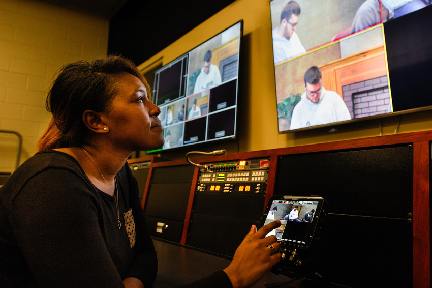 A student in a video production studio monitoring multiple screens and operating a touchscreen tablet, focused on live footage displays.