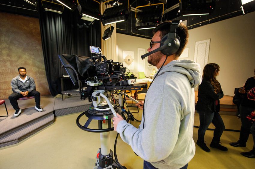 A cameraman operating professional video equipment in a studio, with a person seated on a set in the background.