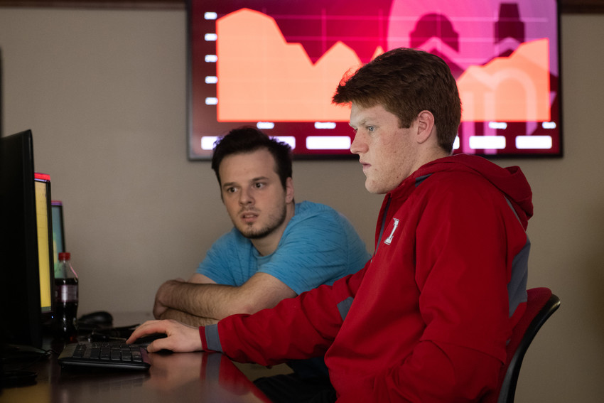 Two students working on a computer in a classroom, with a large screen displaying a red graph in the background.
