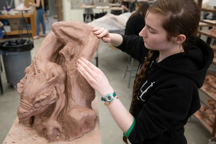 A dedicated art student in a black hoodie meticulously sculpts a large, intricate clay dragon, focusing on the fine details of its face and scales. The scene is set in a busy ceramics studio, filled with tools and other works in progress, highlighting the creative and hands-on environment of the workshop.