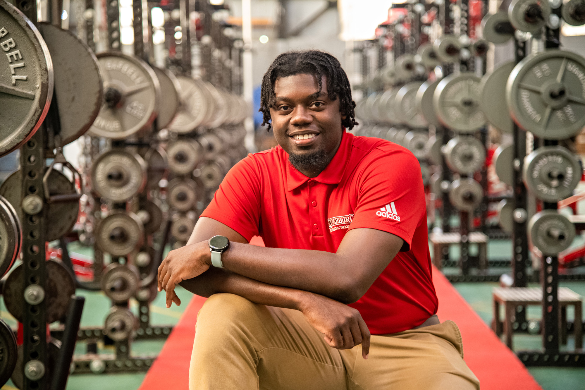 A smiling black man with short locs, kneeling with his arms resting on one knee. He is surrounded by racks of dumbell weights.