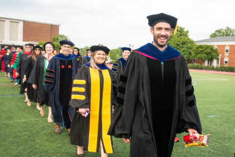 Faculty members dressed in academic regalia participate in a graduation procession on a green field, smiling and walking in a line, with buildings and trees in the background on a sunny day.