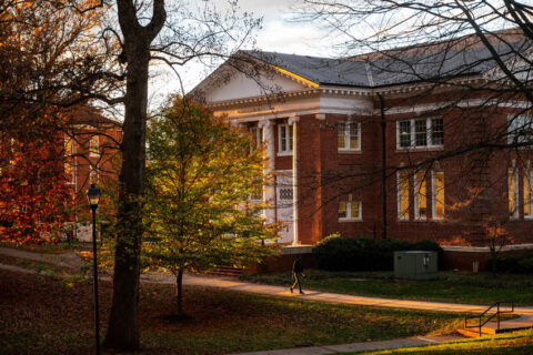 A historic brick building with white columns is illuminated by golden sunlight during the fall season. Trees with autumn foliage surround the building, and a person walks along the pathway, adding a serene and picturesque atmosphere.