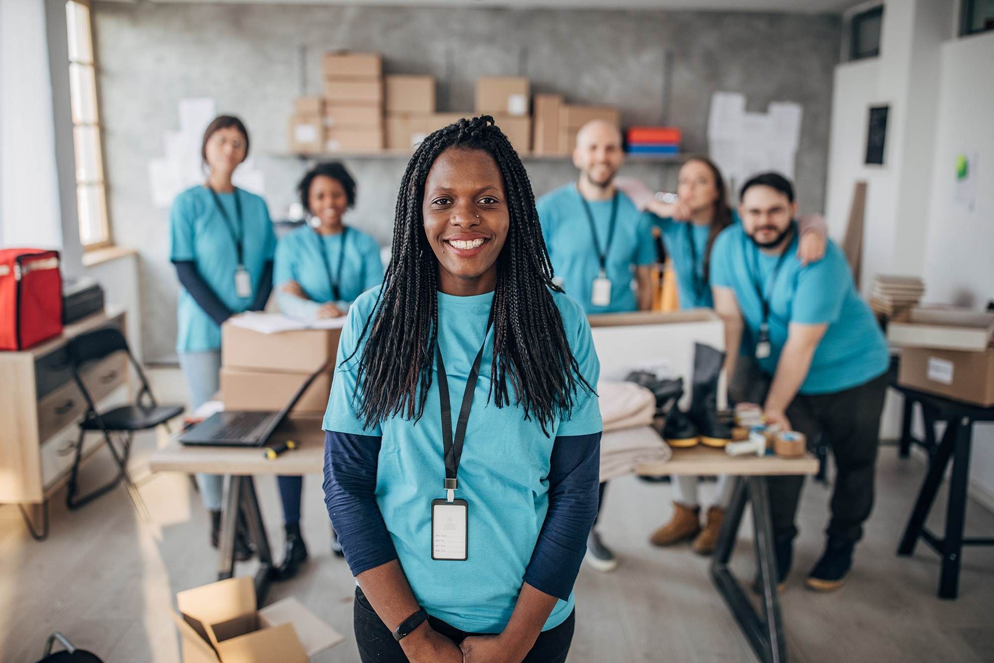 A smiling black woman with long braids, stands in front of a mixed group. They are all wearing matching light blue shirts and lanyards.