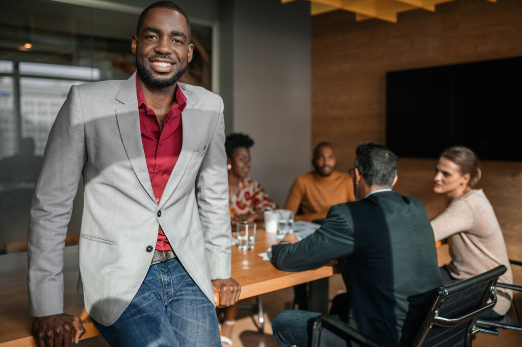 A smiling man leans against a conference room table. He is wearing a red shirt and a gray jacket. Behind him sit a group of four people having a discussion at the conference table.