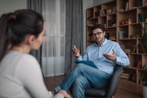 A man wearing glasses and a blue button down shirt sits in a chair and gestures as he speaks to the woman across from him. He has a clipboard on his lap.