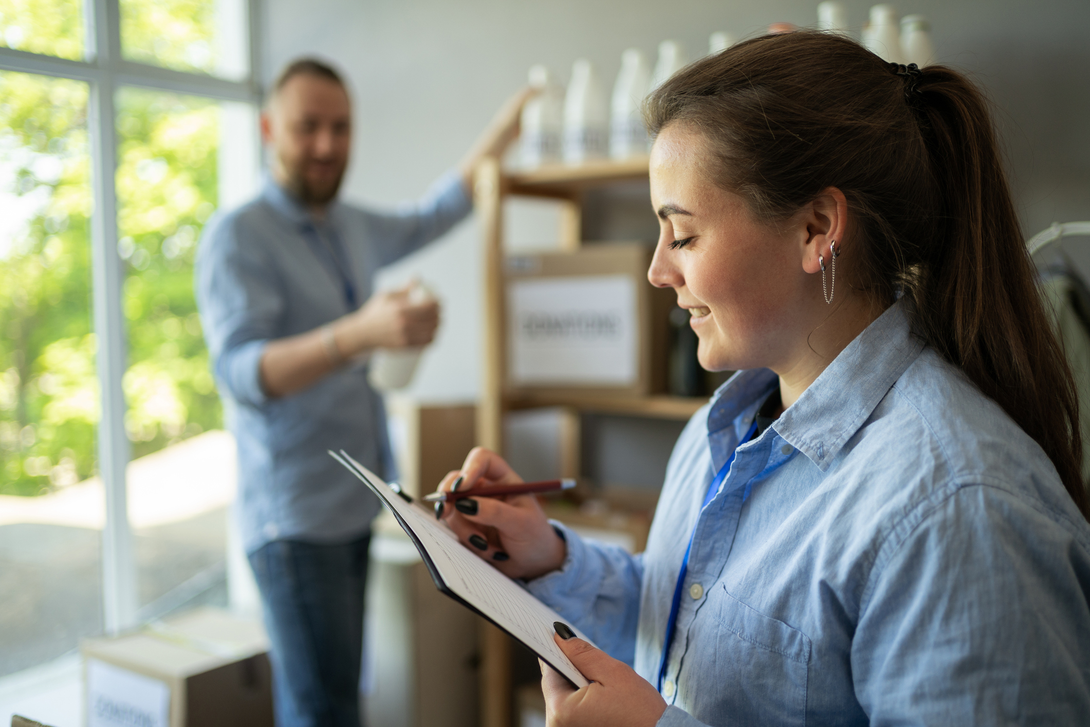 A woman with dark hair pulled into a pony tail reviews information on a clipboard. A man in the background stocks shelves.