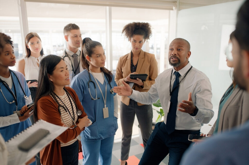 A large and diverse group of hospital staff gathered around a bald man wearing a button down shirt and lanyard.