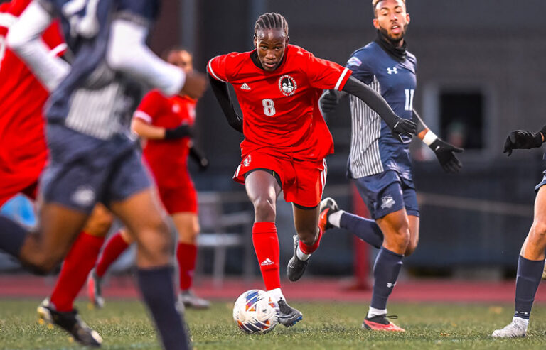 Soccer players in action during a competitive match on a field, with a player in a red jersey (number 8) dribbling the ball skillfully while being pursued by opponents in blue jerseys. The image captures the intensity and focus of the players mid-game.