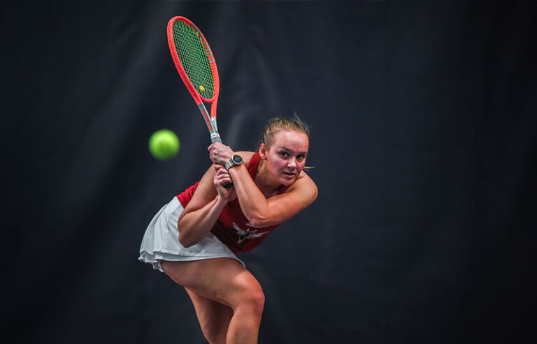 A tennis player in action, wearing a red top and white skirt, preparing to return a shot with a focused expression. The bright green tennis ball is captured mid-air against a dark background, emphasizing the intensity of the moment.