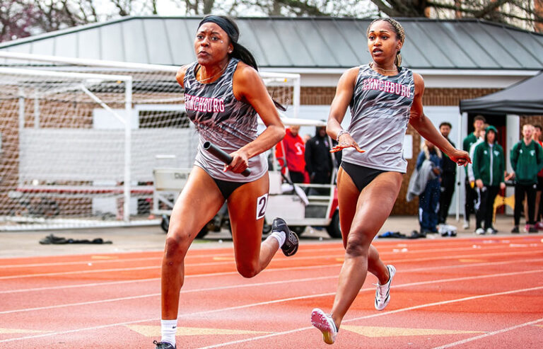 A track and field athlete mid-competition, captured during a hurdle race. The athlete demonstrates focus and agility while clearing a hurdle, with an outdoor track and spectators visible in the background.