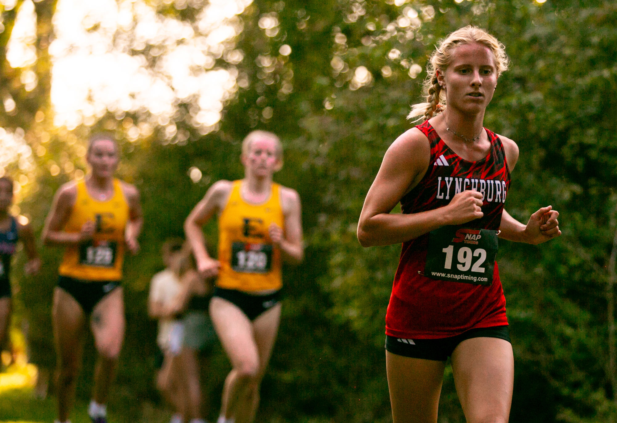 A focused Lynchburg athlete, number 192, competes in a cross-country race through a wooded area, with competitors trailing behind her amidst a backdrop of lush greenery and dappled sunlight.