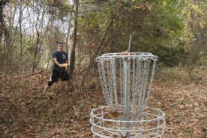 A man in a dark t-shirt and shorts confidently throws a yellow disc towards a disc golf basket, surrounded by dense woods, demonstrating skill and enjoyment in a casual disc golf game.