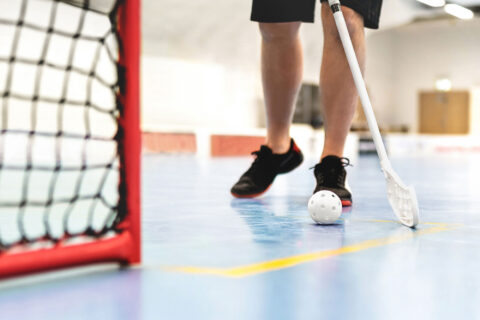 A player with a floor hockey stick poised near a ball on a smooth indoor court, with the goal net visible in the frame.