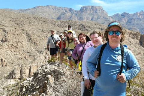 A group of people sitting on rocky terrain during a hiking trip, with a mountainous backdrop. They appear to be enjoying the outdoor adventure, wearing casual and hiking gear, and smiling at the camera.