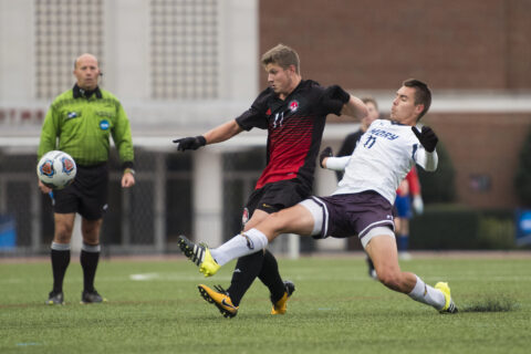 A dramatic moment in a men's soccer game with two players in mid-action; one in a black jersey extends his leg to control the ball while the other in white attempts a tackle, under the watchful eye of a referee in the background.
