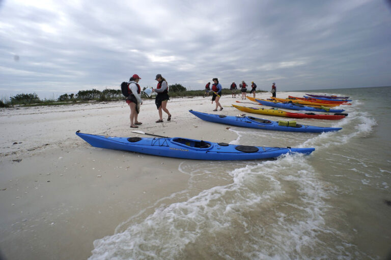 Group of people wearing life vests standing on a sandy beach with colorful kayaks lined up along the shoreline, set against a cloudy sky and gentle waves.