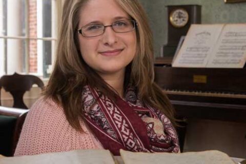 A woman with glasses and long blonde hair sits in a historic room, smiling at the camera. She wears a pink sweater and a patterned scarf. An open antique book is in the foreground, and a piano with sheet music is visible in the background.
